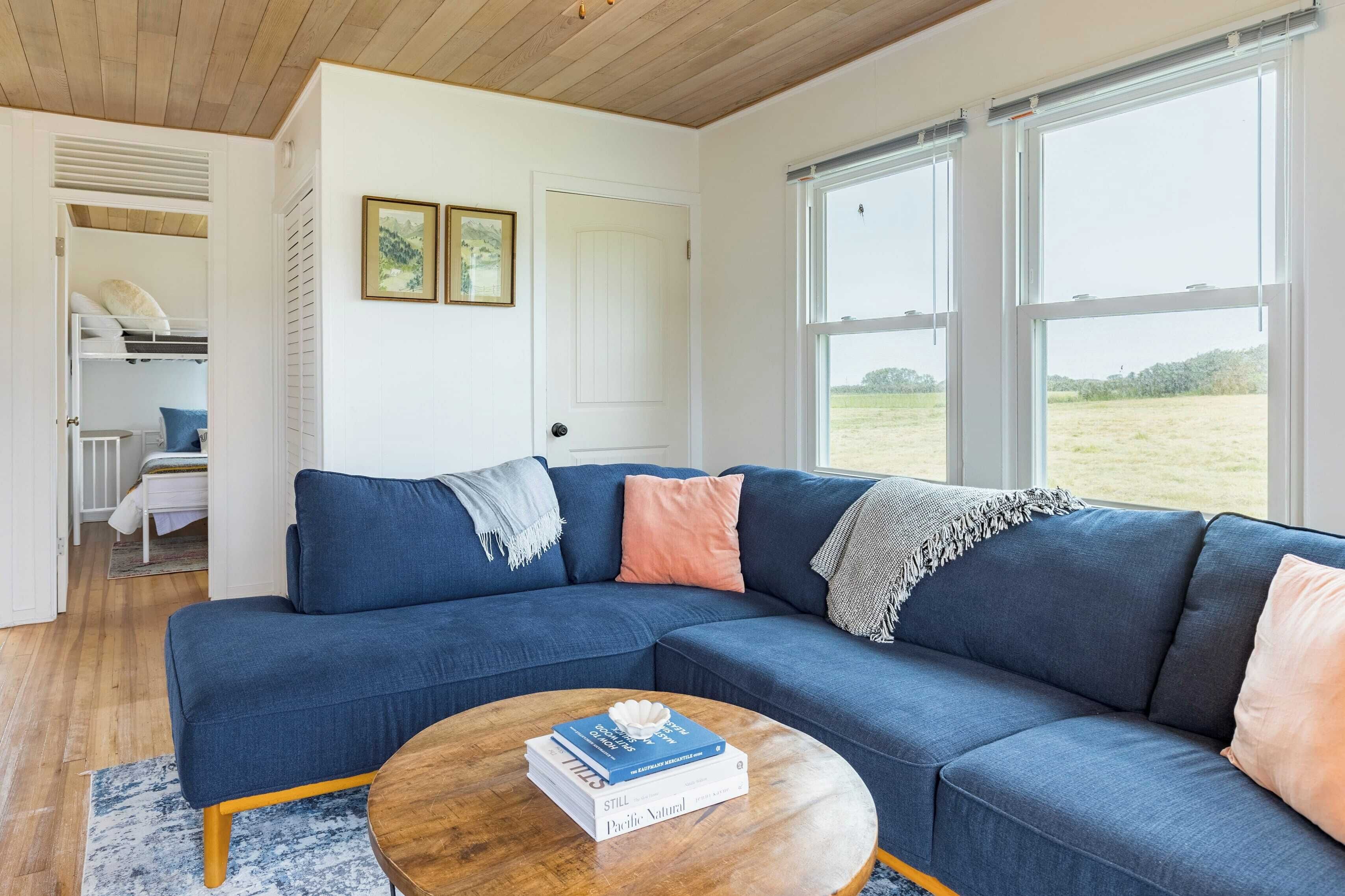 A modern living room with a blue sectional sofa, a wooden coffee table, and a view of a grassy field through large windows.