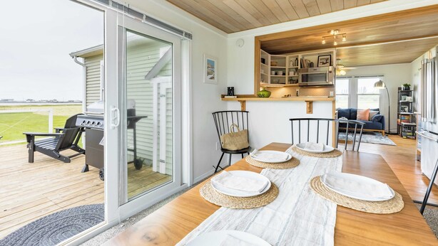 A spacious deck with a sliding glass door leading to a patio area, a barbecue grill, and a dining table set with plates and placemats. - Swan Lake Cottage by Avantstay Views + Firepit (Oak Harbor)