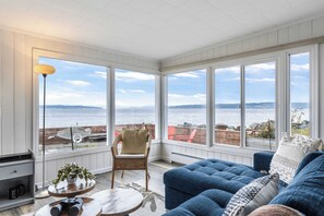 A sunroom with large windows overlooking a body of water, furnished with a blue sofa, wicker chair, and a small round table. - Harrington Hideaway by Avantstay Beach Access (Coupeville)