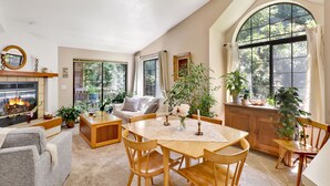 Un salon spacieux doté d’une cheminée, un coin repas avec des meubles en bois et de grandes fenêtres offrant une vue sur la verdure.