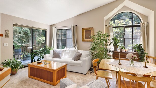 Un salon spacieux avec un canapé blanc, une table basse en bois et un coin repas comprenant une table et des chaises. De grandes fenêtres laissent entrer beaucoup de lumière naturelle et offrent une vue sur la verdure environnante.