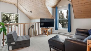 A modern living room with a wooden ceiling, a large flat-screen TV, a black leather sofa, and a view of trees through a window. - Amazing location with private hot tub, sauna, laundry and ground level suite.  (Silver Star Mountain)