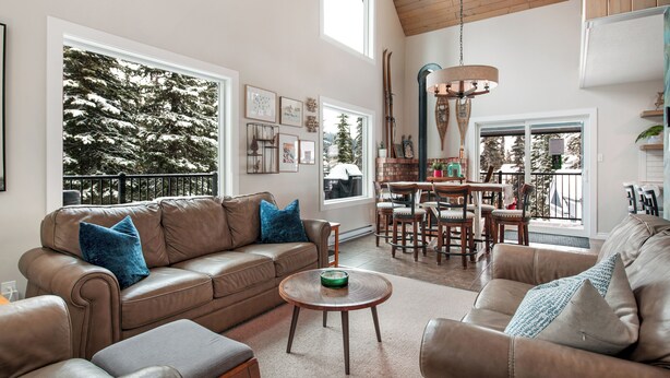 A spacious living room with leather sofas, a dining area, and large windows offering a view of trees. - Amazing location with private hot tub, sauna, laundry and ground level suite.  (Silver Star Mountain)