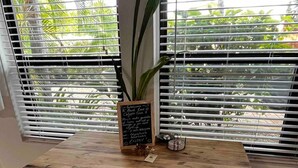 Una mesa de madera con cuatro taburetes, una planta en maceta y un cartel en una pizarra en una habitación con ventanas grandes y persianas blancas.