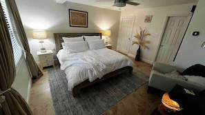 Un dormitorio con cama, lámparas de noche, mesita de noche, una silla, un florero con plantas secas y una puerta.