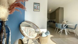 A modern living room with a blue accent wall, a wicker chair, a dining area with white chairs, and a kitchen in the background.