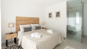 Una habitación de hotel moderna con cabecera de madera, cabina de ducha de vidrio y cama con ropa de cama blanca y almohadas estampadas.
