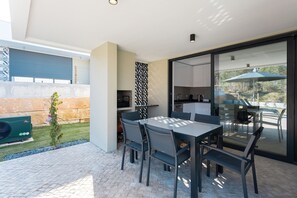 Uma área moderna de refeições ao ar livre com mesa, cadeiras, churrasqueira e vista para a cozinha através de uma porta de vidro.