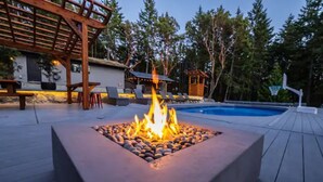 Ein quadratisches Feuerbecken aus Steinen, umgeben von einer Holzterrasse, in der Nähe eines Pools und einer Sitzgruppe im Freien.