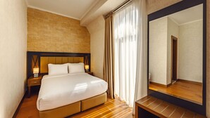A hotel room with a large bed, a nightstand with a lamp, a wooden bench, a large mirror, and a brick-patterned wall. - Chateau Kvirike (Kobuleti)
