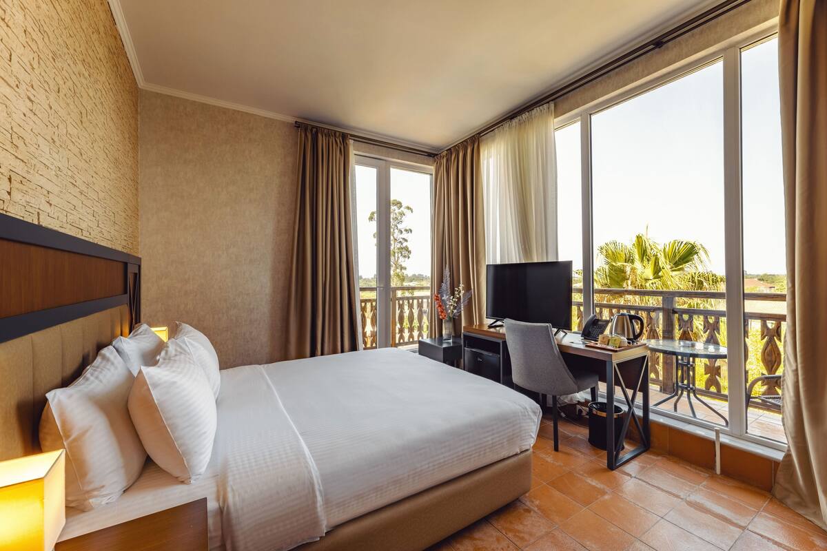 A hotel room with a large bed, a desk with a computer, and a TV. There is a balcony with a view of palm trees.