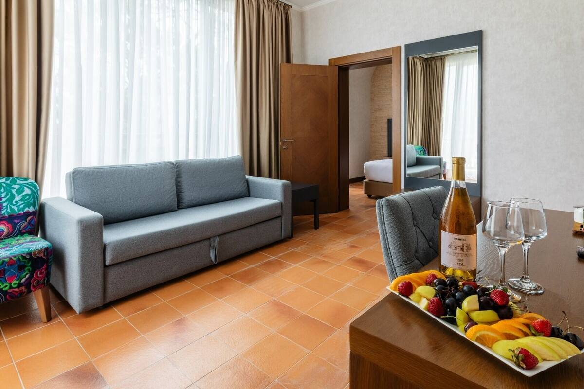 A hotel room with a grey sofa, a small table with a fruit platter and wine glasses, and a mirror on the wall.
