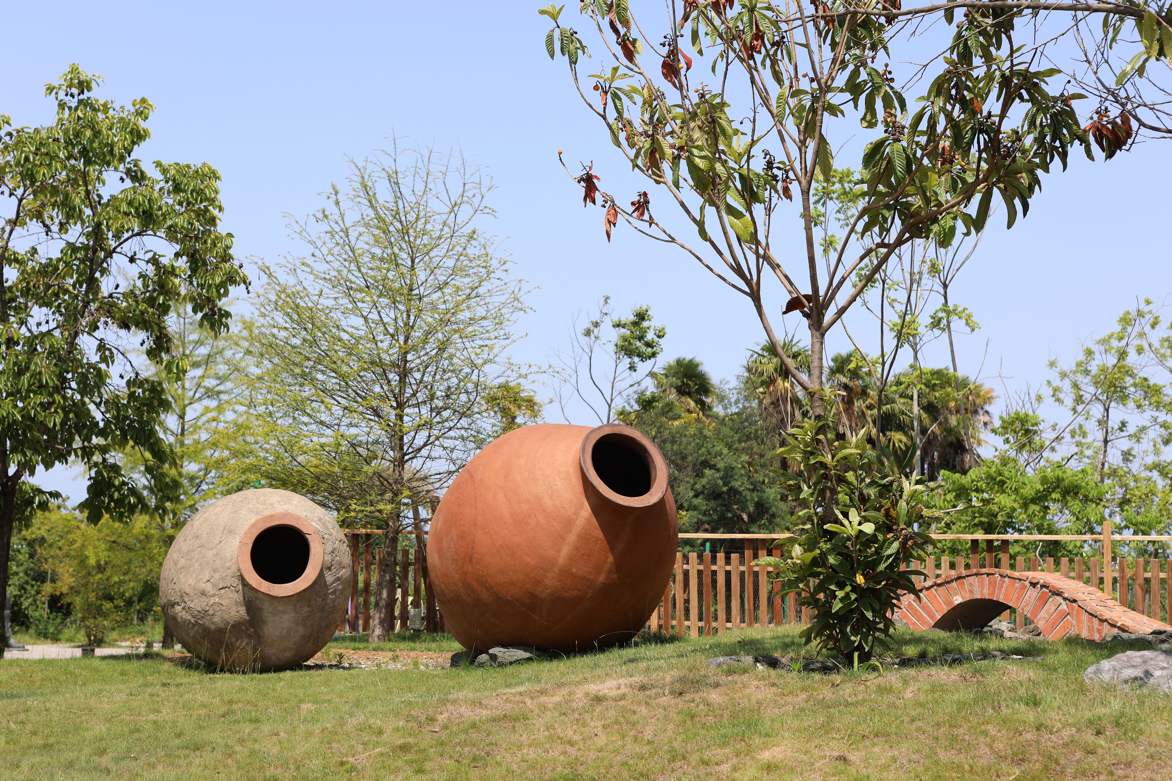 A large clay pot and a smaller pot in a grassy area with trees and a brick bridge in the background.