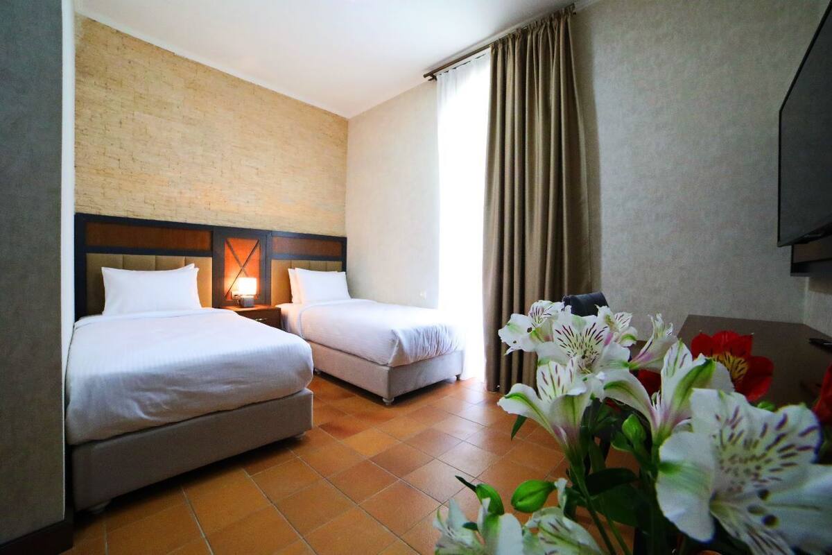 A hotel room with two beds, a brick accent wall, a large window with curtains, and a flat-screen TV.
