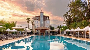 A swimming pool with lounge chairs and umbrellas, a building with a balcony, and palm trees. - Chateau Kvirike (Kobuleti)
