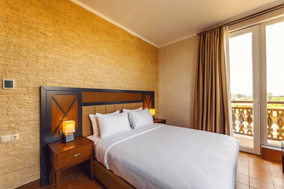 A hotel room with a brick wall, a wooden headboard, a large bed with white linens, two bedside lamps, and a sliding glass door leading to a balcony.