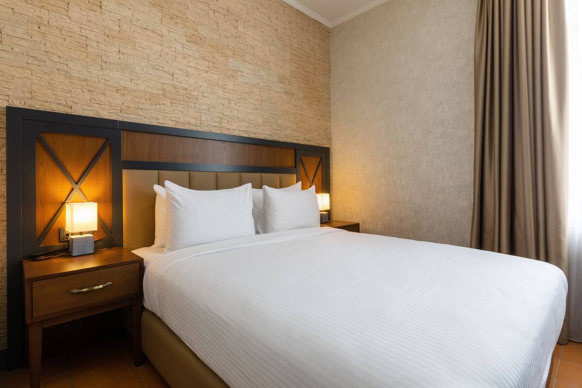 A hotel room with a brick-patterned wall, a wooden headboard, a large bed with white linens, two bedside tables with lamps, and a window with curtains.