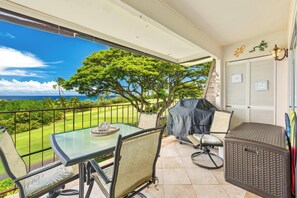 A balcony with a glass table, chairs, a covered grill, and a view of a green landscape and ocean. - Keauhou Akahi Ocean View Condo Overlooking Kona Country Club Golf Course (Kailua-Kona)