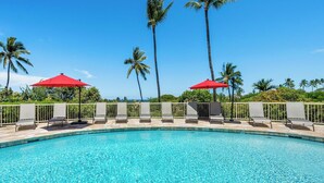 A pool with clear blue water, surrounded by lounge chairs and red umbrellas, with palm trees in the background.