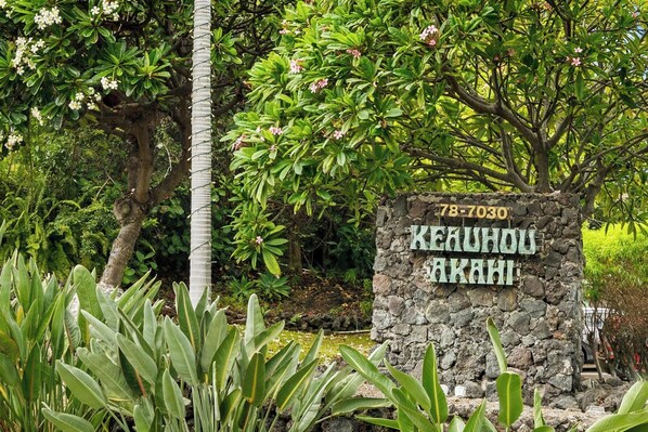 Um letreiro de pedra com 'KEAUHOU AKAHI' e o número '78-7030' em um jardim exuberante.