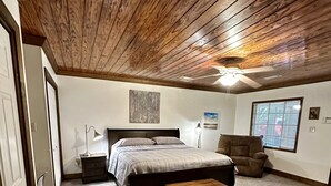 Une chambre avec un plafond en bois, un lit avec une banquette, une chaise, une table de chevet et une fenêtre avec des stores.