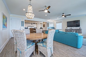 A spacious living room with a large round wooden table surrounded by wicker chairs with blue floral cushions, a teal sofa, and a chandelier.