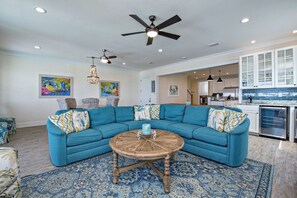 A living room with a blue sectional sofa, a round wooden coffee table, and a patterned rug. There are ceiling fans, pendant lights, and artwork on the walls. - BeachFront 18Bed Pool EasyBreezy (Gulf Shores)