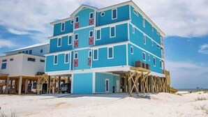 A multi-story building with a blue exterior, a covered entrance, and a sandy beach in the background. - BeachFront 18Bed Pool EasyBreezy (Gulf Shores)