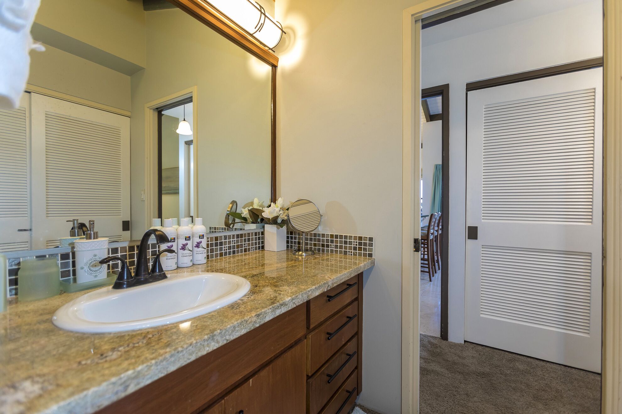 Un baño con encimera de granito, un lavabo blanco y un mueble con cajones de madera. Hay artículos de aseo en la encimera, y un espejo sobre el lavabo.