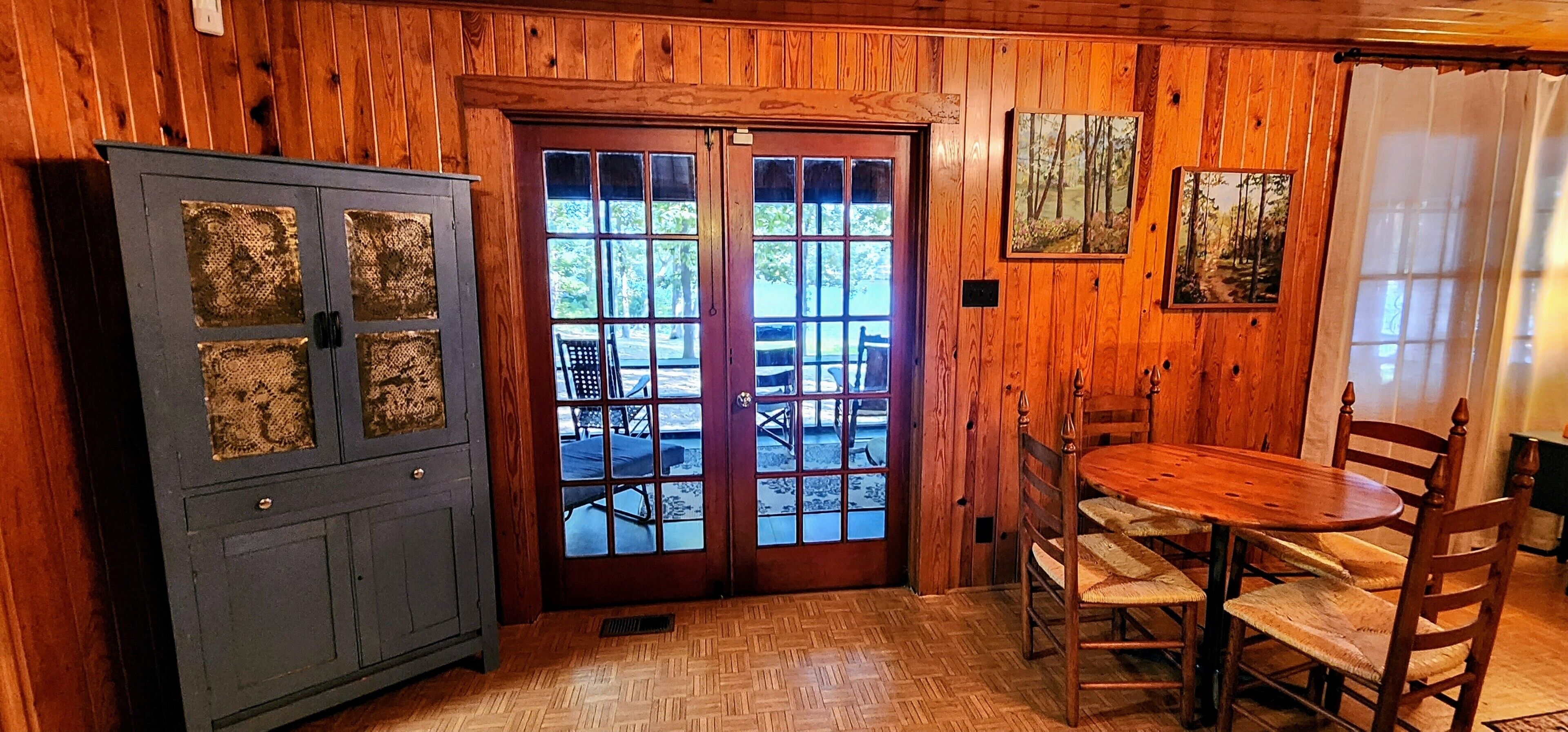 A wooden interior with a dining table and chairs, a cabinet with decorative panels, and a door leading outside.