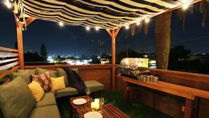 Une terrasse extérieure dotée d’un auvent à rayures, une table en bois dressée pour le repas, un barbecue et un canapé avec des coussins.
