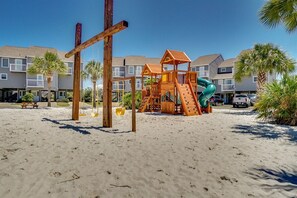 Un terrain de jeux au bord de la plage, avec un toboggan en bois et des structures d’escalade, entouré de palmiers et bordé d’une rangée de maisons de ville.