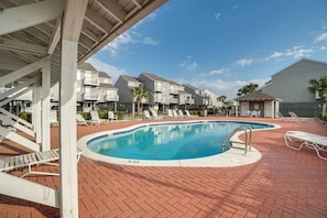 A swimming pool with lounge chairs, a covered area, and residential buildings in the background.