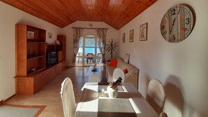 A cozy living room with a wooden ceiling, a television on a wooden stand, a dining table with chairs, a sofa with cushions, and a wall clock. - Sponga 2 Apartment with balcony and sea view (Polače)