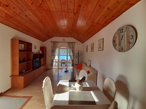 Un salon chaleureux avec un plafond en bois, un téléviseur posé sur un meuble en bois, une table à manger avec des chaises, un canapé avec des coussins et une horloge murale.