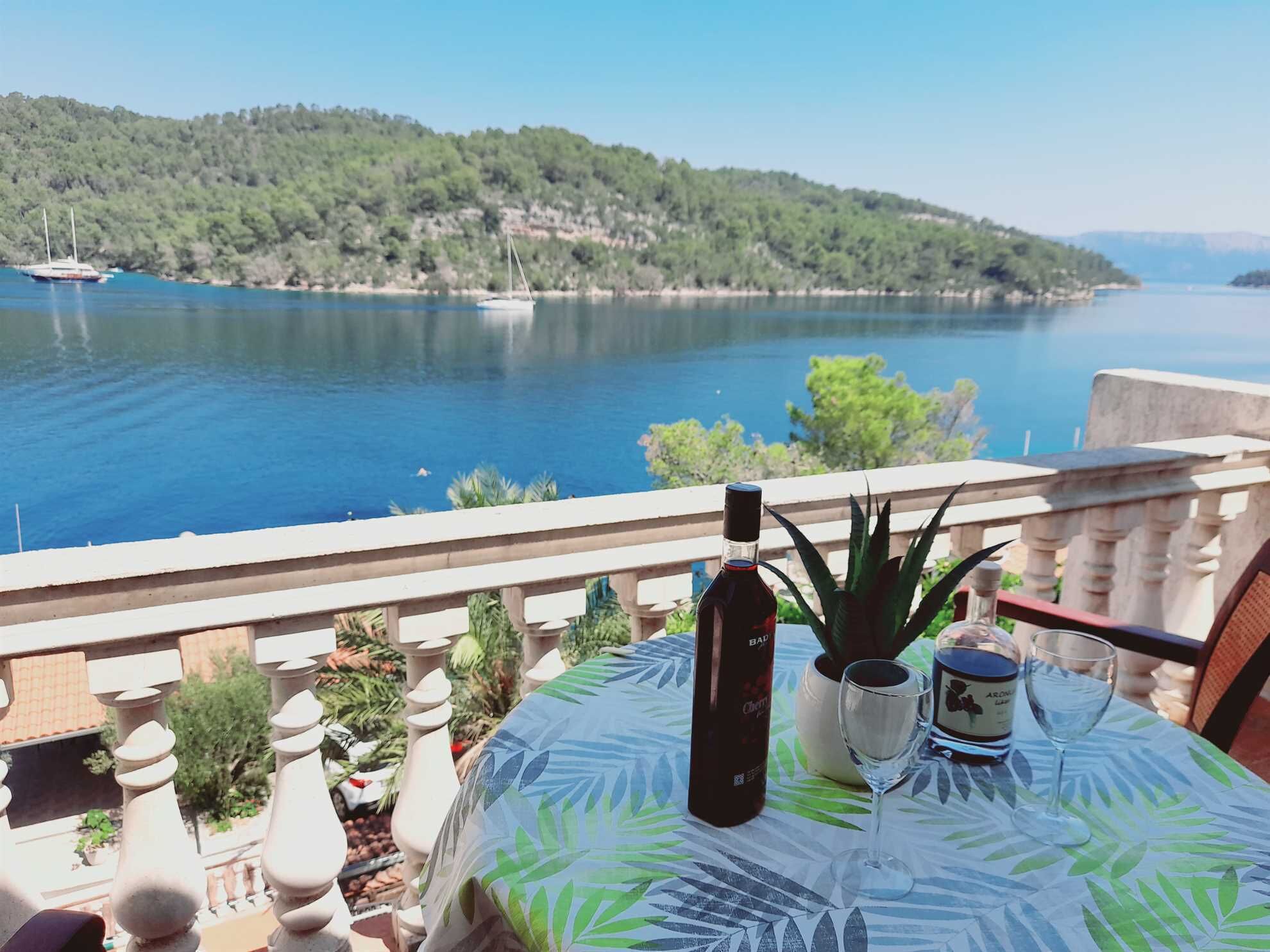 A balcony with a table set for two, overlooking a lake with boats and a forested hillside.