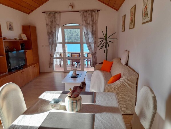 A cozy living room with a sofa, a coffee table, and a television on a wooden cabinet. There is a window with curtains, a plant, and a view of the outside.