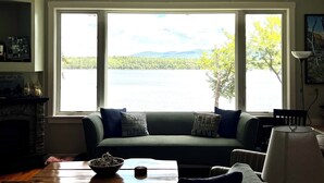A living room with a large window overlooking a lake, a green sofa with cushions, a fireplace, and a table with a bowl.