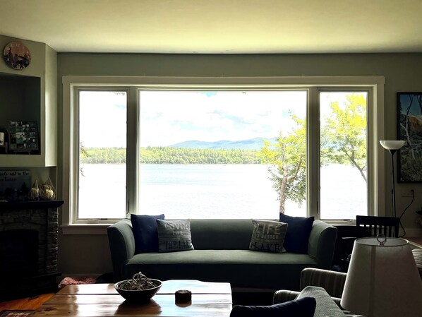 Un salon doté d’une grande fenêtre donnant sur un lac, d’un canapé vert agrémenté de coussins, d’une cheminée et d’une table sur laquelle se trouve un saladier.
