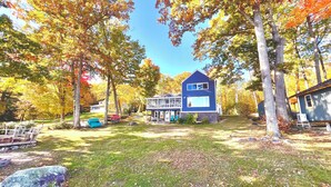 A two-story house with a large deck and a covered porch, surrounded by trees with autumn foliage.