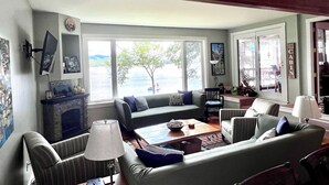 A cozy living room with a fireplace, comfortable seating, and large windows offering a view of the outdoors.