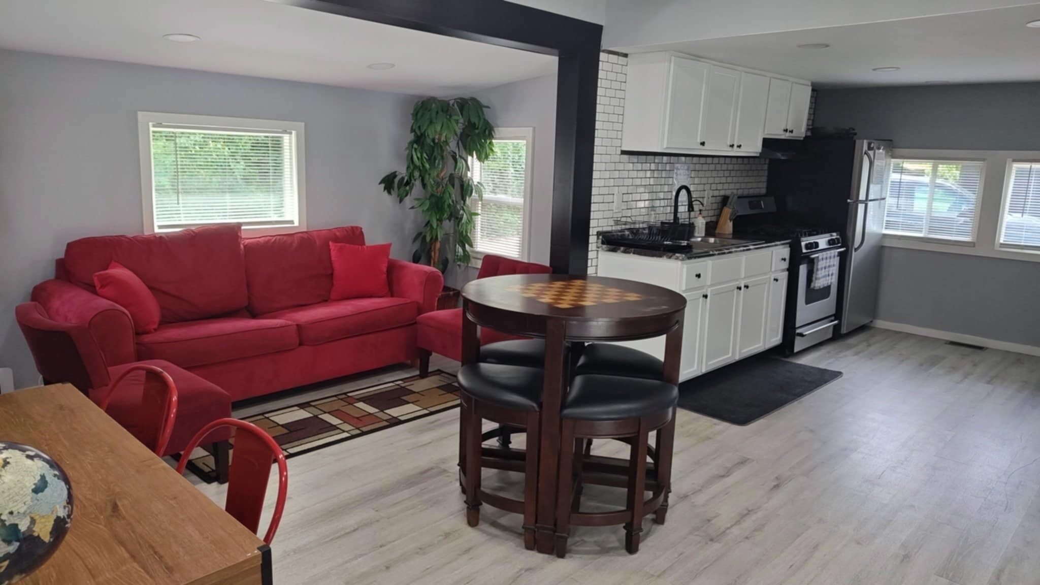 A living room with a red sofa, a wooden dining table with two chairs, a kitchen with white cabinets, a black refrigerator, and a large window with blinds.