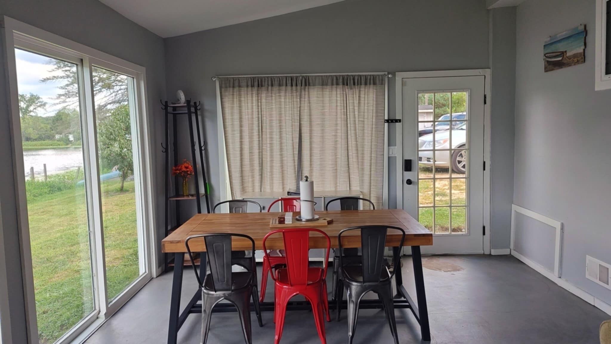Un coin repas avec une table en bois, des chaises noires et rouges, un vase de fleurs et une vue sur un lac, visible à travers de grandes fenêtres.