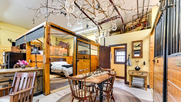 A rustic room with a wooden dining table, chairs, and a large window. - Unique Finger Lakes Converted Horse Barn w/ Patio! (Geneva)