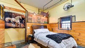 A wooden-paneled room with a bed, a window, a fan, and a wall with a horse painting and bags of California beans. - Unique Finger Lakes Converted Horse Barn w/ Patio! (Geneva)