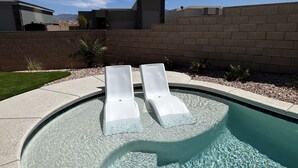Une piscine avec deux chaises longues blanches, de l’eau cristalline, un mur de briques et un ciel dégagé.