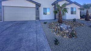 Une maison moderne à deux étages, dotée d’un garage, d’une allée et d’un jardin aménagé avec des rochers et des palmiers.