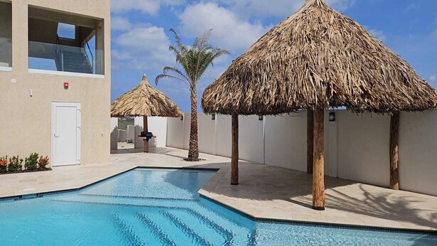 A swimming pool with thatched umbrellas, a white building, and a palm tree. - Gloednieuw Appartement met Zeezicht. Fantastisch Gelegen 5 min van the Beach (Oranjestad)