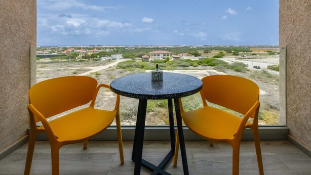 Un balcon avec deux chaises jaunes et une petite table ronde, d’où l’on peut admirer un paysage lointain composé de bâtiments et de verdure.