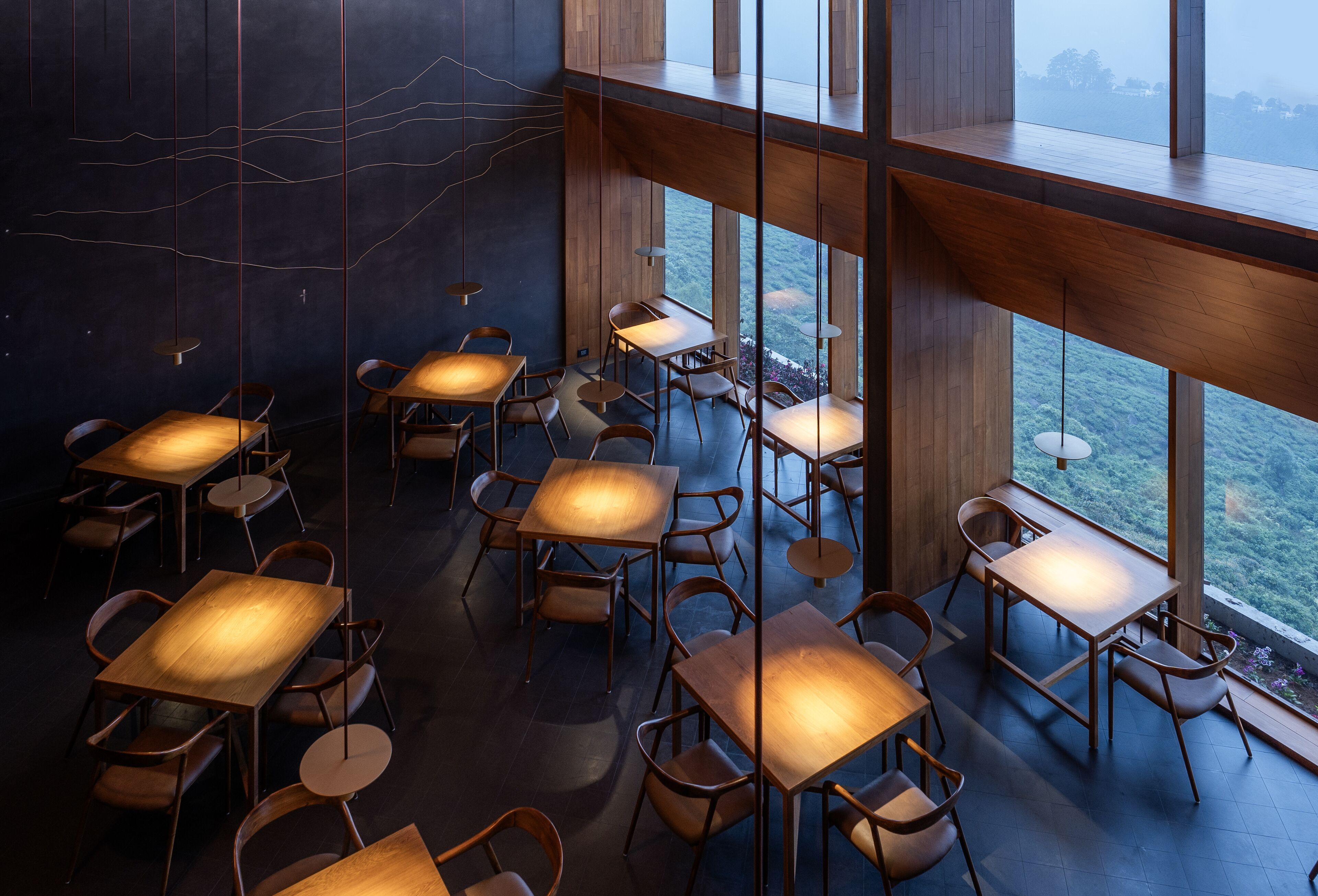 A modern interior with multiple wooden tables and chairs arranged in a spacious layout, large windows offering a view of the outdoors.
